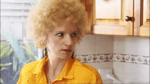 a woman with blonde curly hair is wearing a yellow shirt and earrings in a kitchen .