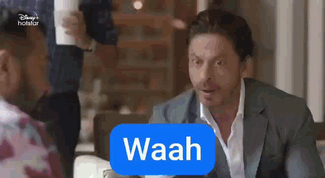 a man in a suit and white shirt is sitting at a table with a sign that says waah .