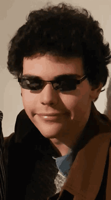 a young man wearing sunglasses and a brown coat smiles for the camera