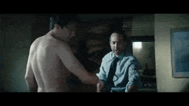 a shirtless man in a blue shirt and tie stands next to another shirtless man in front of a painting ..