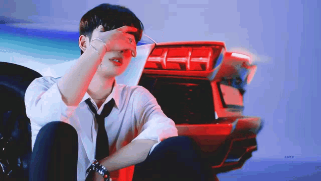 a man in a white shirt and tie is sitting in front of a red car covering his face with his hand .