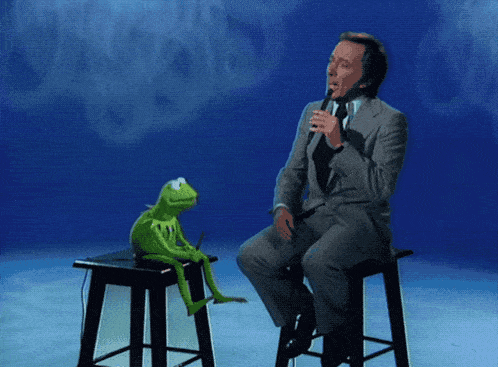 a kermit the frog sits on a stool next to a man singing into a microphone