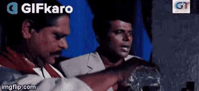 two men are standing next to each other in a dark room and one of them is holding a glass .
