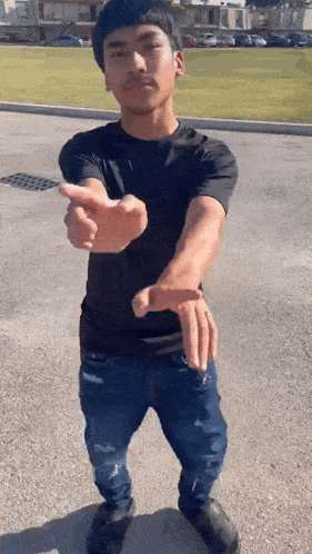 a young man in a black shirt and blue jeans is standing in a parking lot and pointing at the camera .