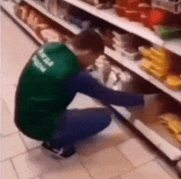 a man in a green vest is kneeling down in front of a shelf in a grocery store .