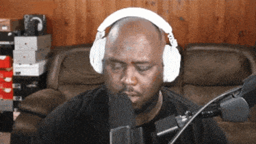 a bald man wearing white headphones stands in front of a microphone with his eyes closed