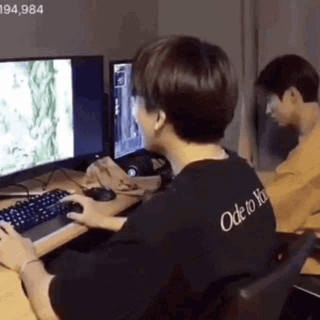 two men are playing a video game on a computer . one of the men is wearing a shirt that says ode to you .