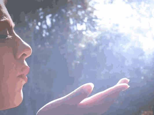 a close up of a person blowing a kiss with the sun shining through the trees in the background