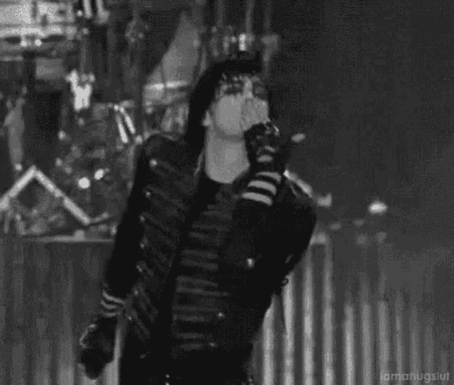 a black and white photo of a man in a black jacket singing into a microphone on a stage .