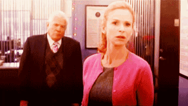 a woman in a pink cardigan stands in front of a man in a suit