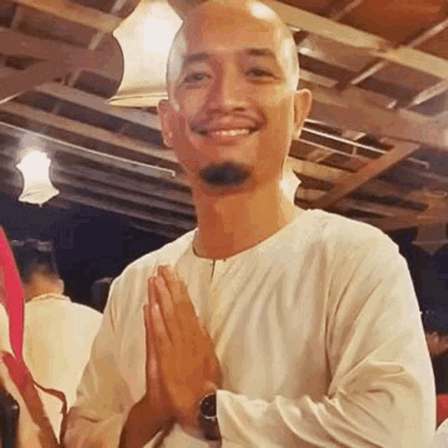 a bald man with a beard is smiling with his hands folded in prayer .