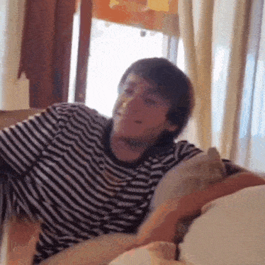 a man in a striped shirt is sitting on a couch in front of a window and smiling .