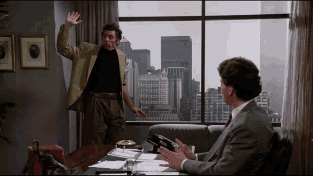 a man in a suit stands in front of a window while another man sits in front of a desk