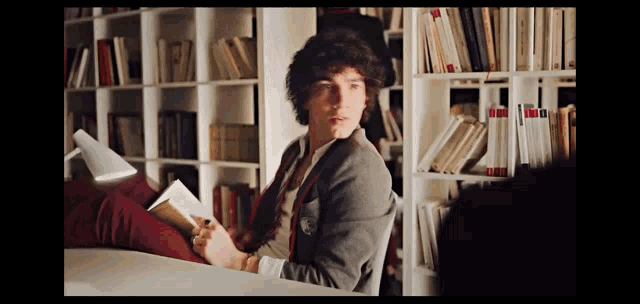 a young man is sitting in front of a bookshelf reading a book ..