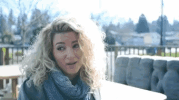 a woman with blonde curly hair wearing a scarf and a sweater is sitting at a table .