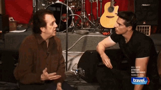 two men are talking in front of a drum set and a guitar that says ' travel ' on the bottom