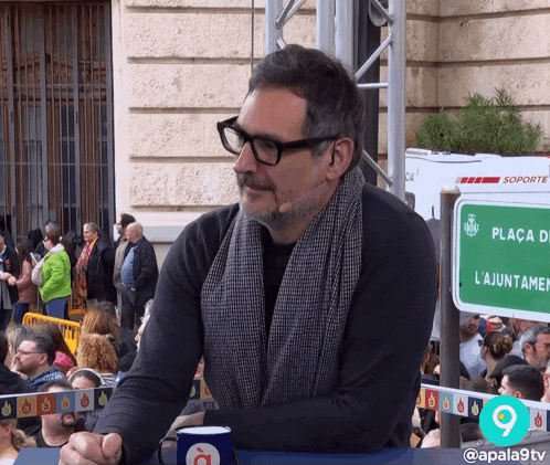 a man wearing glasses sits in front of a sign that says plaça de l'ajuntament