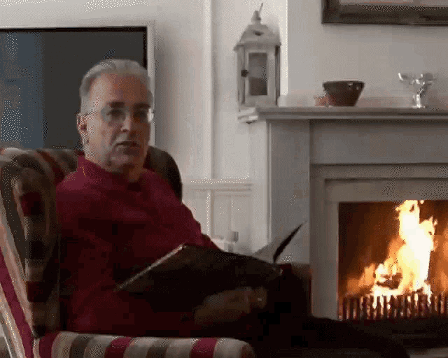 a man in a red sweater is sitting in front of a fireplace reading a book
