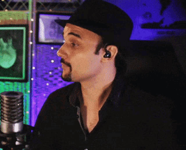 a man with a beard wearing a hat and ear buds stands in front of a microphone
