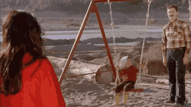 a girl in a red cape is looking at a child on a swing