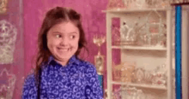 a little girl in a blue shirt is smiling in a room with a pink wall .