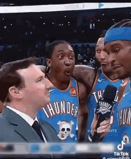 a man in a suit and tie is talking into a microphone while basketball players are standing around him .