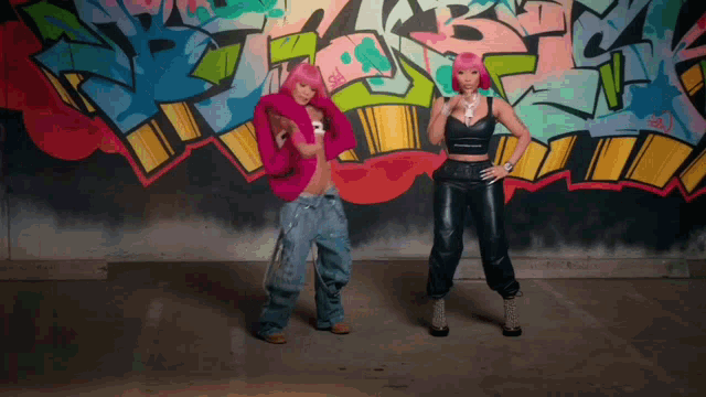 two women are dancing in front of a wall with graffiti on it including the letter s