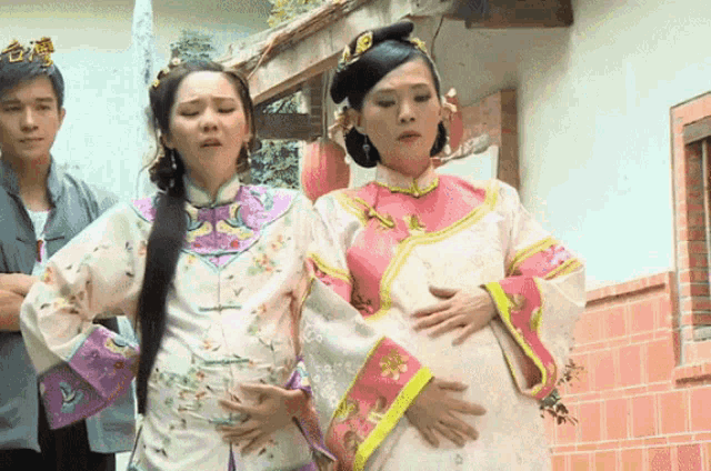 a woman in a pink and white dress holds her belly while a man stands behind her
