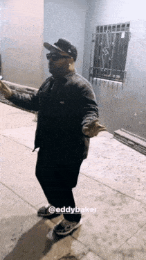 a man wearing sunglasses and a hat is standing on a sidewalk with the name eddybaker on the bottom right