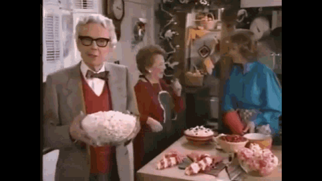 a man in a suit and bow tie is holding a bowl of popcorn while standing in a kitchen .