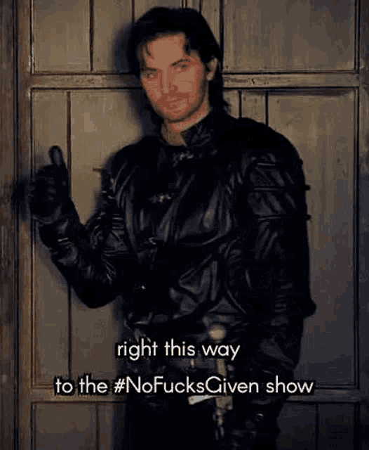 a man in a leather jacket is standing in front of a wooden door giving a thumbs up .