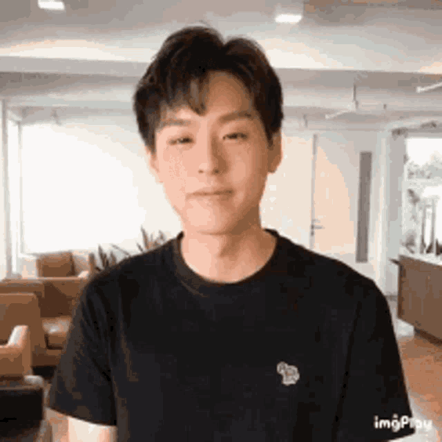 a young man wearing a black t-shirt is standing in a living room looking at the camera .