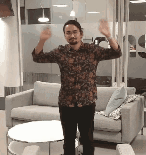 a man in a floral shirt stands in front of a couch with his arms outstretched