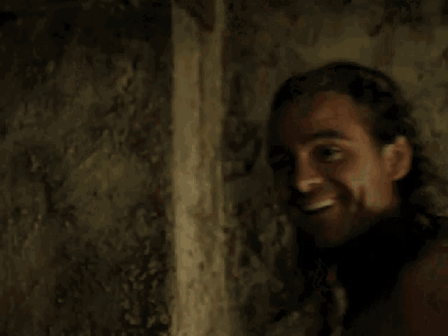 a shirtless man with long hair leaning against a wall and smiling