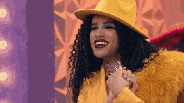 a drag queen wearing a yellow hat and a fur coat is holding her hands together and smiling .