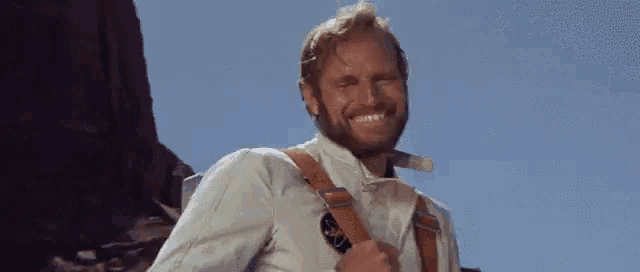 a man with a beard wearing a white shirt and brown straps smiles
