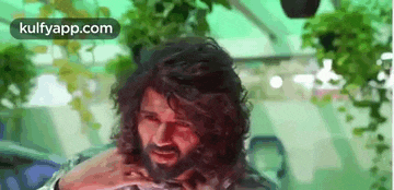 a man with long curly hair and a beard is sitting in a greenhouse .