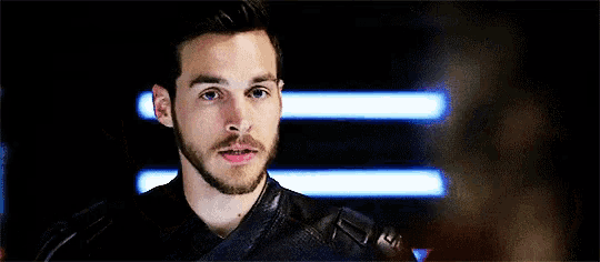 a man with a beard is wearing a black leather jacket and standing in front of a dark background .