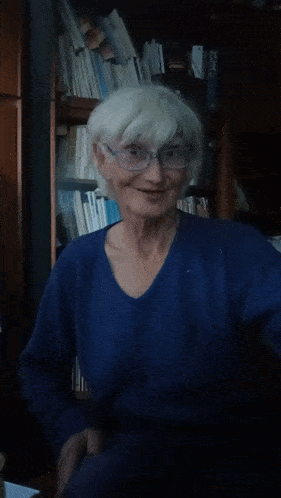 an elderly woman wearing glasses and a blue sweater smiles in front of a bookshelf