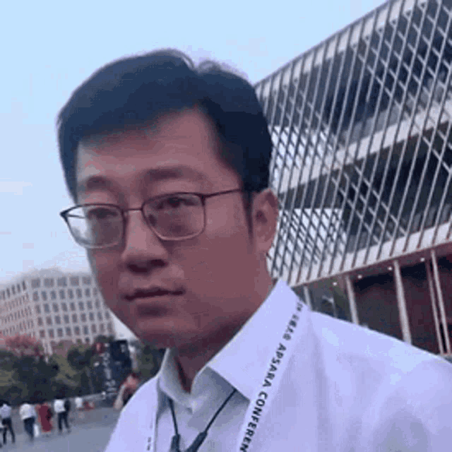 a man wearing glasses and a lanyard with the word aasara on it is standing in front of a large building .