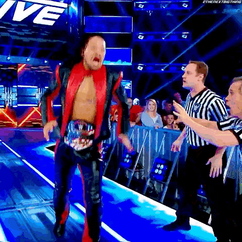 a man in a red and black suit is walking down a wrestling ring while a referee talks to him .