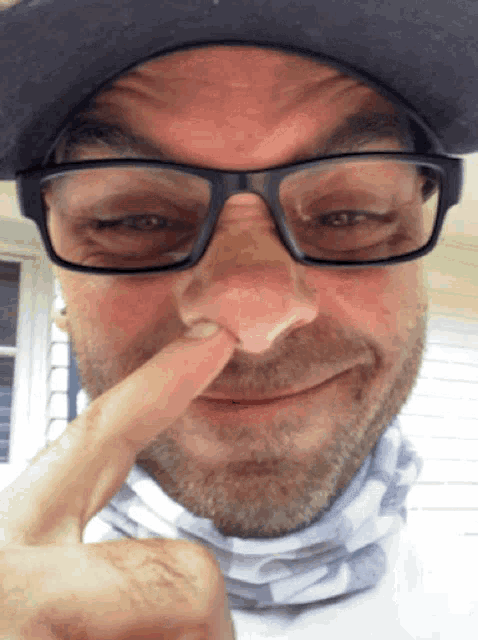 a man wearing glasses and a hat holds his finger up to his nose