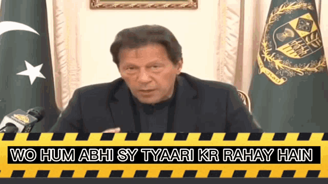 a man in a suit stands in front of a microphone with the words wo hum abhi sy tyaari kr rahay hain above him