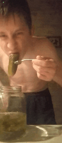 a shirtless man eats a pickle with a spoon