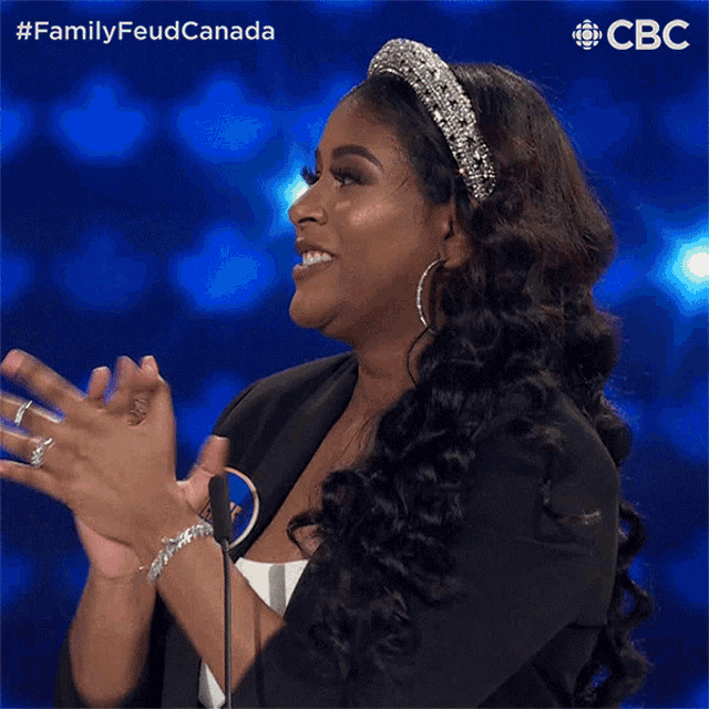 a woman wearing a headband and earrings applauds in front of a microphone with the words family feud canada behind her