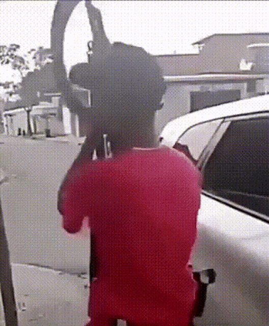 a man in a red shirt is holding a gun in front of a car .