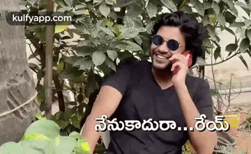 a man wearing sunglasses is talking on a cell phone while sitting in front of a tree .