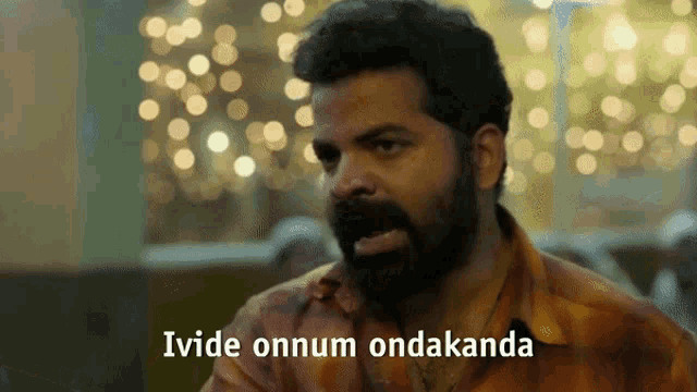 a man with a beard is sitting at a table with the words " ivide onnum ondakanda " written below him