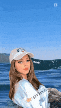 a girl wearing a hat that says us sits on a rock in front of the ocean