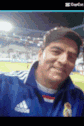 a man in a blue adidas shirt is taking a selfie in front of a soccer field .